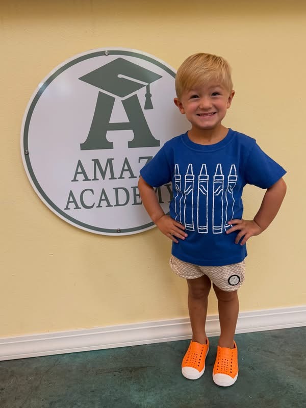A smiling Amaze Academy student in orange shoes standing in front of the school logo